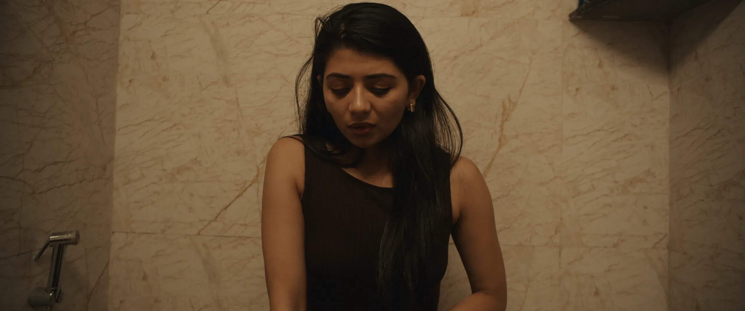 A woman sits against a marble-tiled wall, looking down in warm tungsten light.