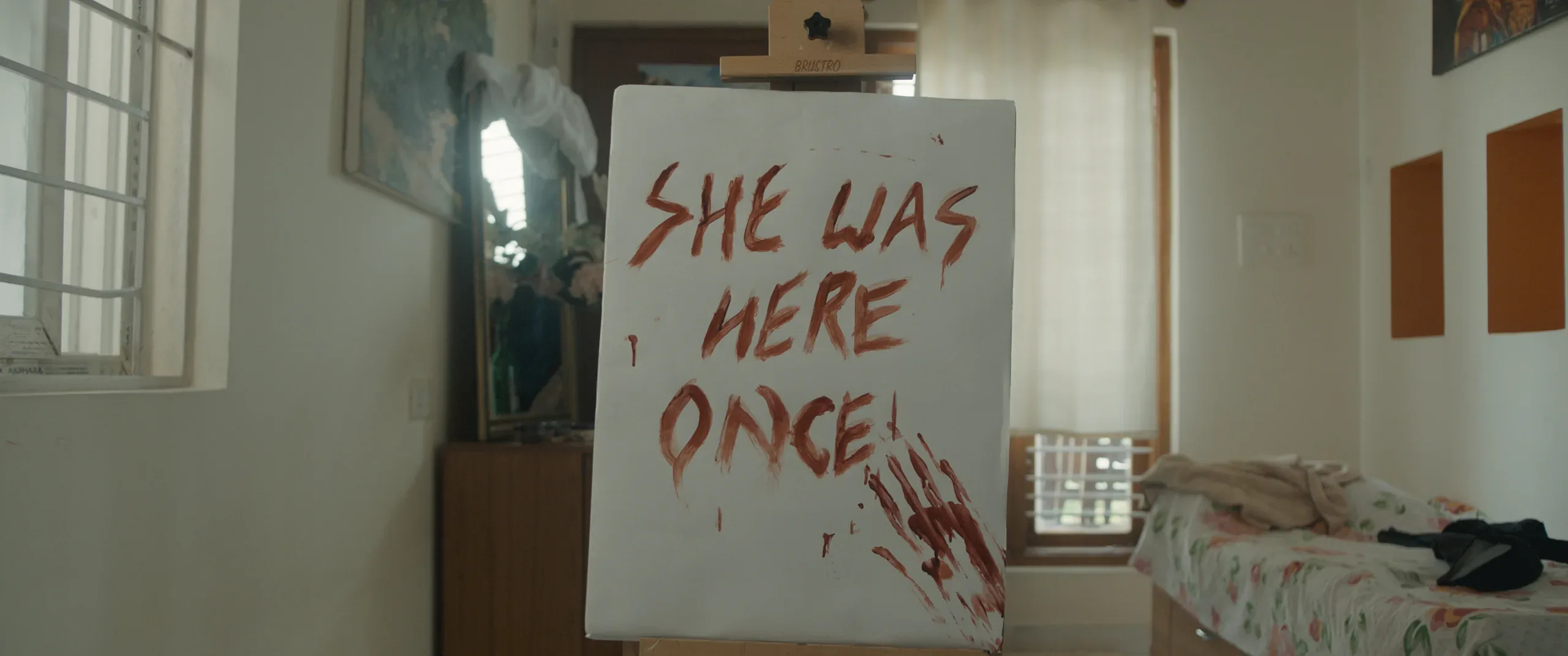 A canvas reads "She was here once" in red beside a bloodied handprint, in a bright bedroom.