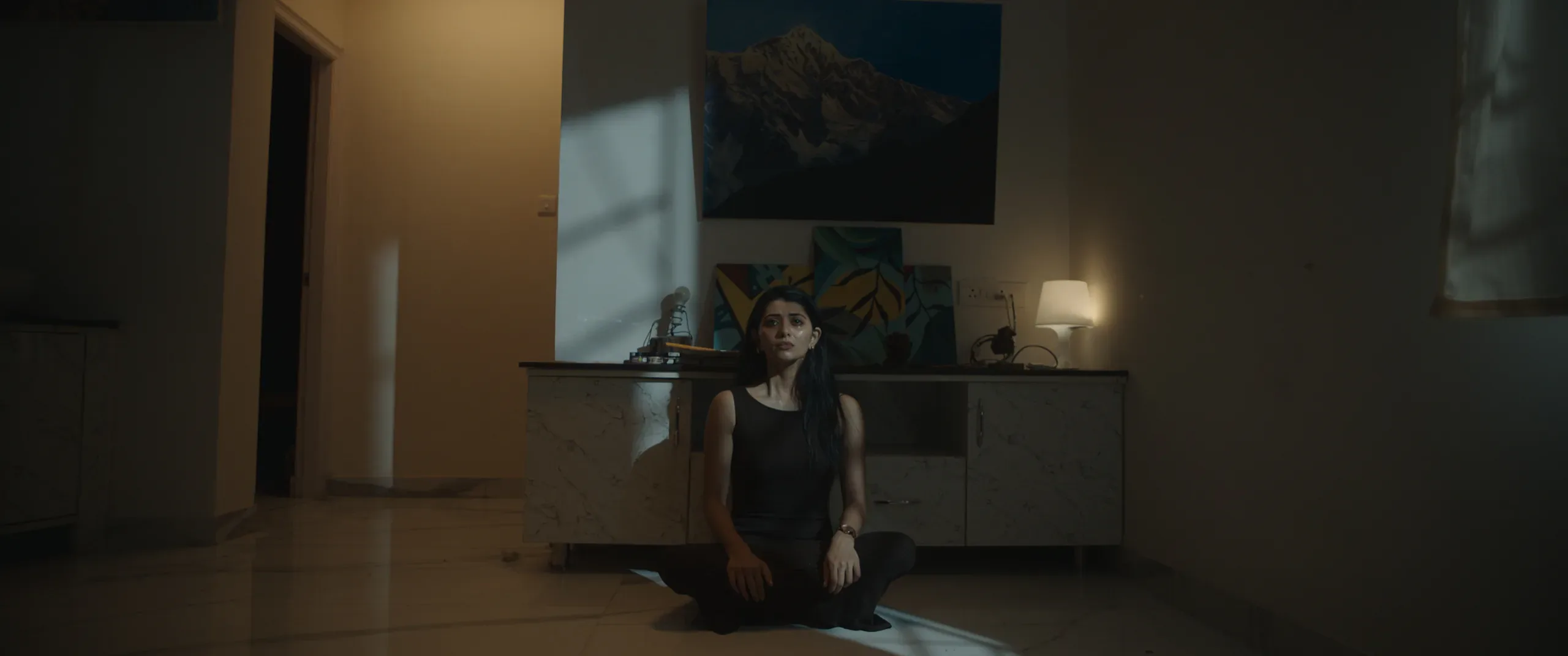 Wide shot of a woman kneeling on the floor of a dim room, a lamp glowing beside framed paintings.