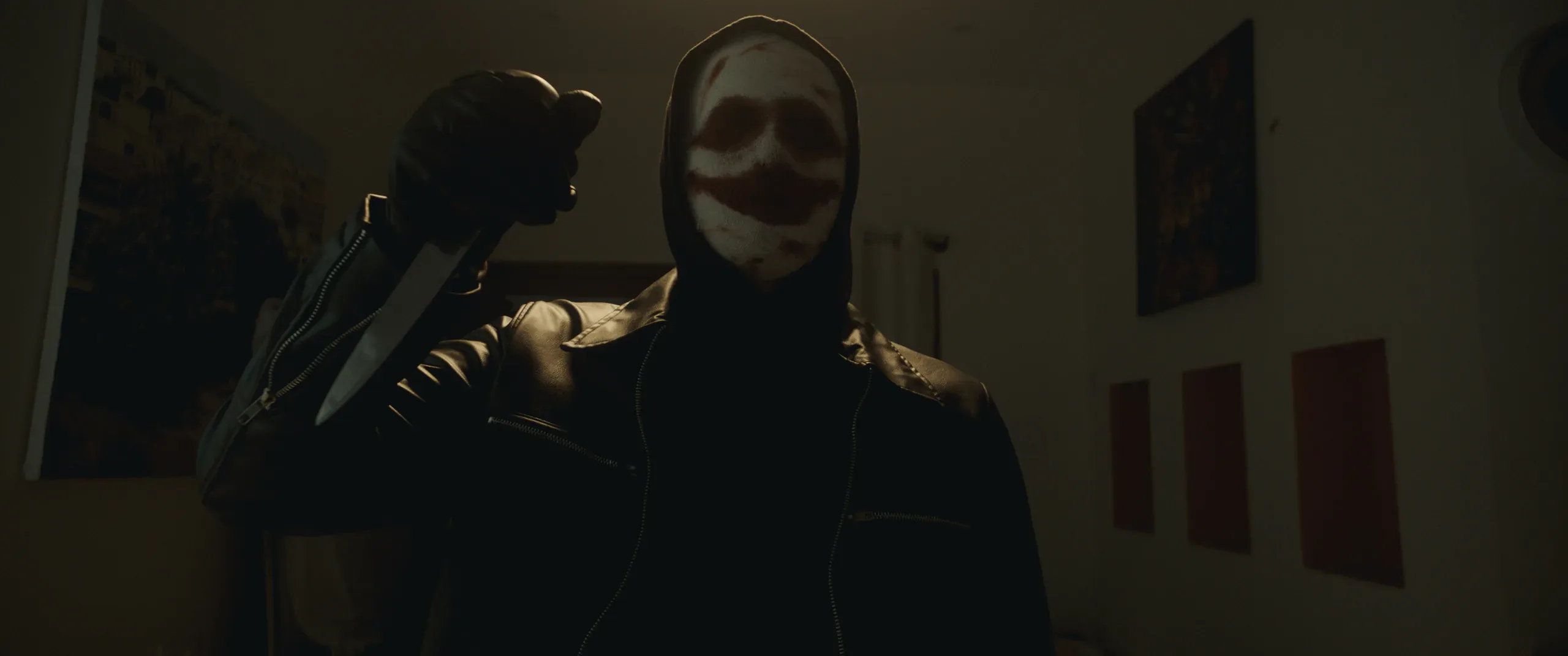 Two shadowy figures, one in a pale mask, stand in a darkened room.